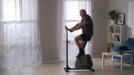 home-training-with-modern-exercise-bike-losing-weight-and-keeping-fit-of-middle-aged-man-wide-shot-in-living-room